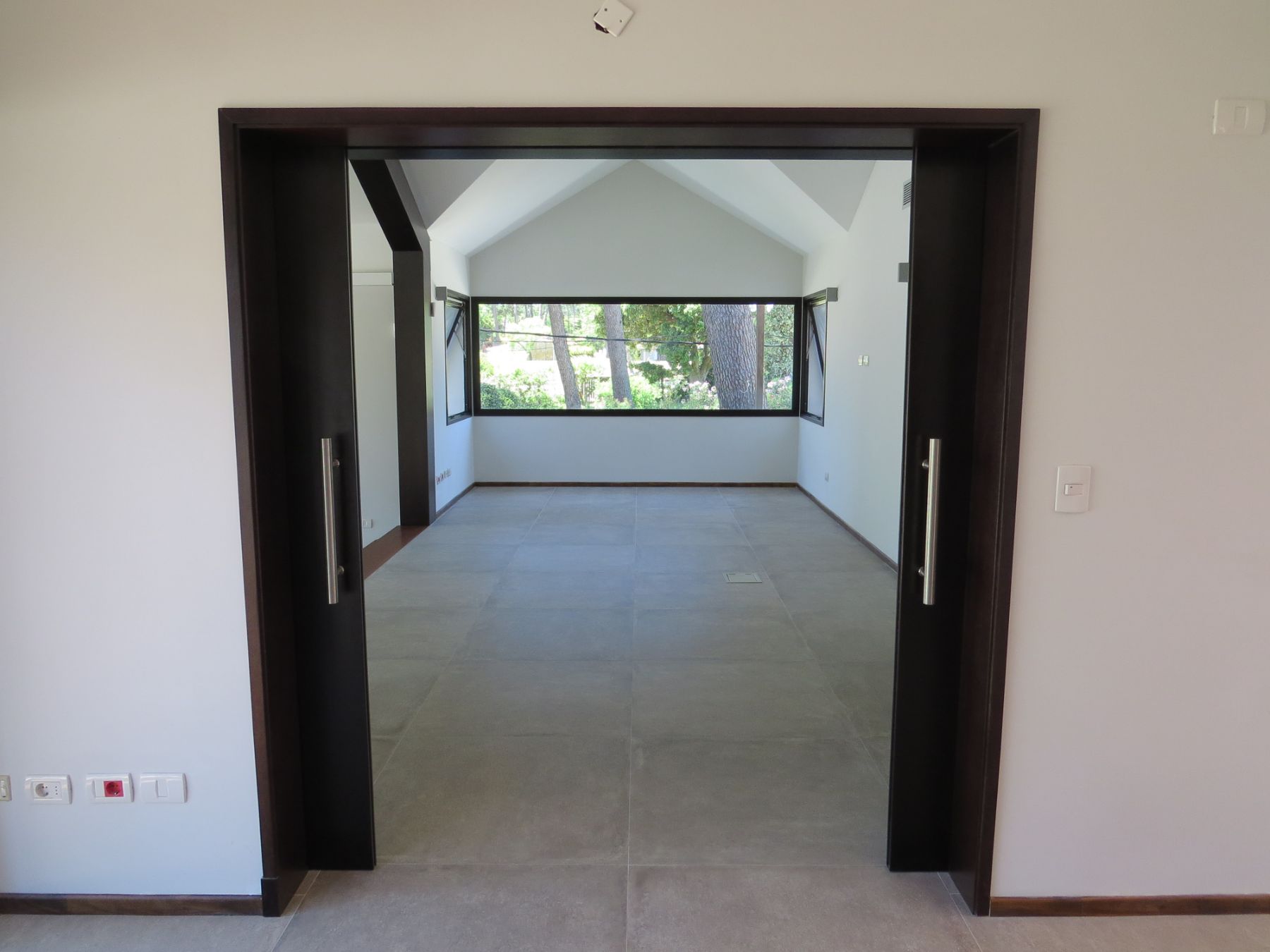 En la imágen se aprecia la puerta que divide la sala principal de la planta alta del sector de escalera y ascensor. Es una puerta corrediza de dos hojas de madera, en el fondo se aprecia una ventana amplia.